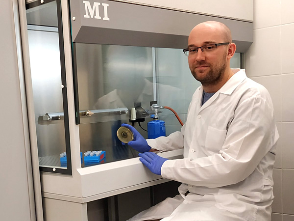 A man in a white coat sits at a fume hood. In his hand he holds a glass dish with a cultivated colony of fungus