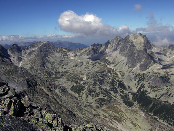 A rocky valley surrounded by high mountains with steep slopes. Above, an almost cloudless blue sky