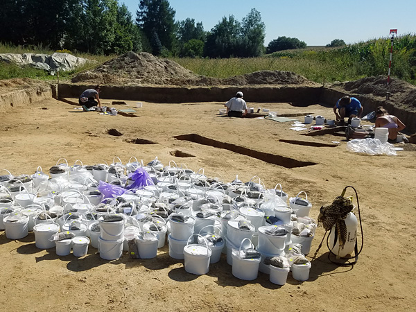 Excavation site: rectangular field without topsoil. In the foreground, several dozen white plastic buckets