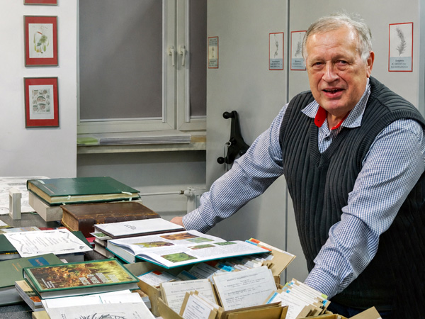 An older man leans on a table covered with books and envelopes with moss specimens; collection cabinets in the background