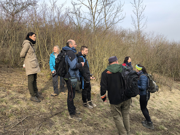 A group of people in field clothes stand on a gentle slope against a background of leafless bushes