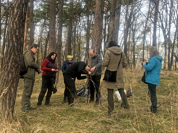 A group of people in field clothes stand among the pine trees around a device mounted on a tripod